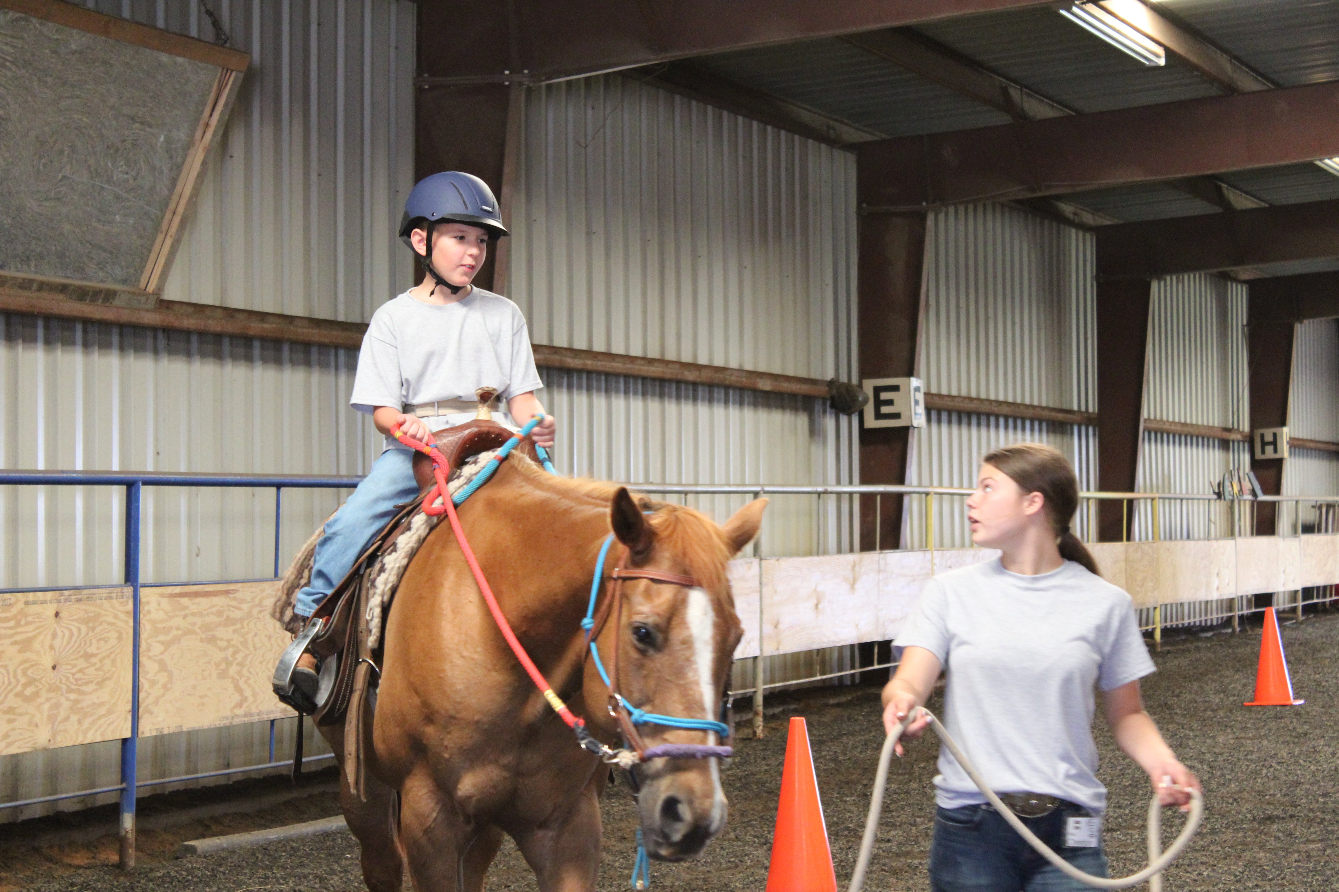 A child sits on the back of a horse, holding the reins, while a volunteer leads the horse around cones in an indoor arena.