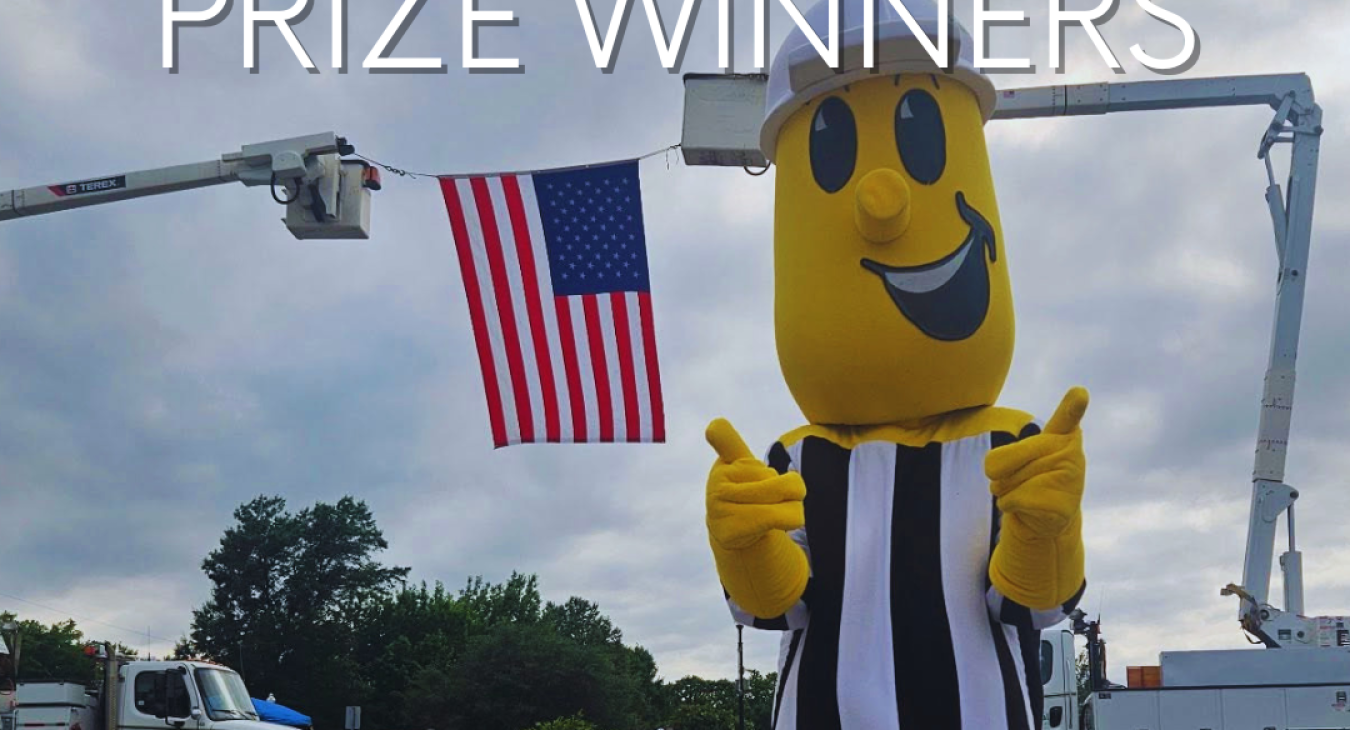 Willie Wiredhand stands in front of the American flag, suspended between two bucket trucks. Test on the image says Annual Meeting Prize Winners.