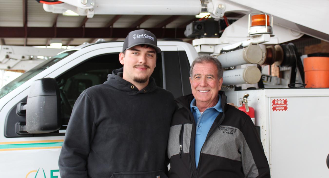 ECE lineman Bryer Crosby, left, and retired ECE Lineman Jack McCullar, right, stand together in front of an East Central bucket truck.