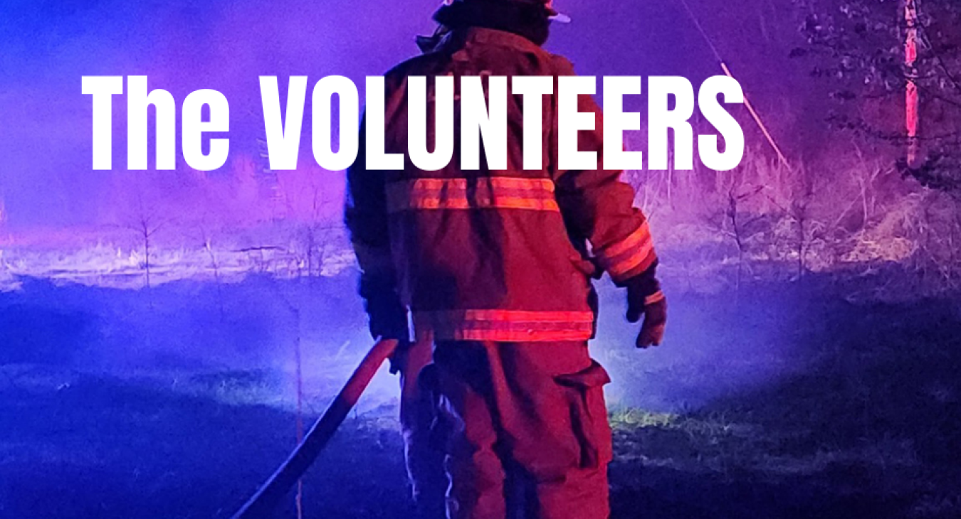 Image of a firefighter spraying with a hose in the dark. The words "The Volunteers" are overlayed in white.